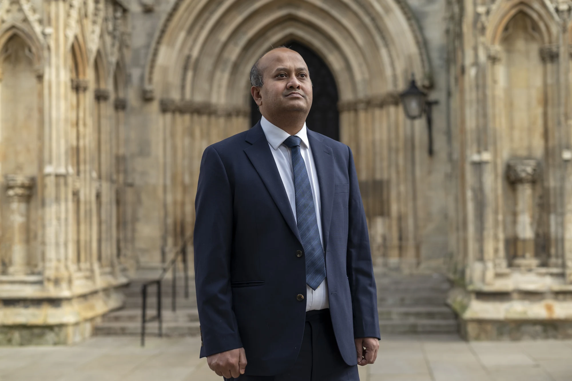 William Gomes in a navy suit beside historic stone architecture